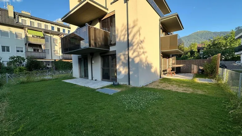 Modernes Gebäude mit Balkonen und einem gepflegten Rasenbereich, umgeben von grüner Landschaft und Bergen.