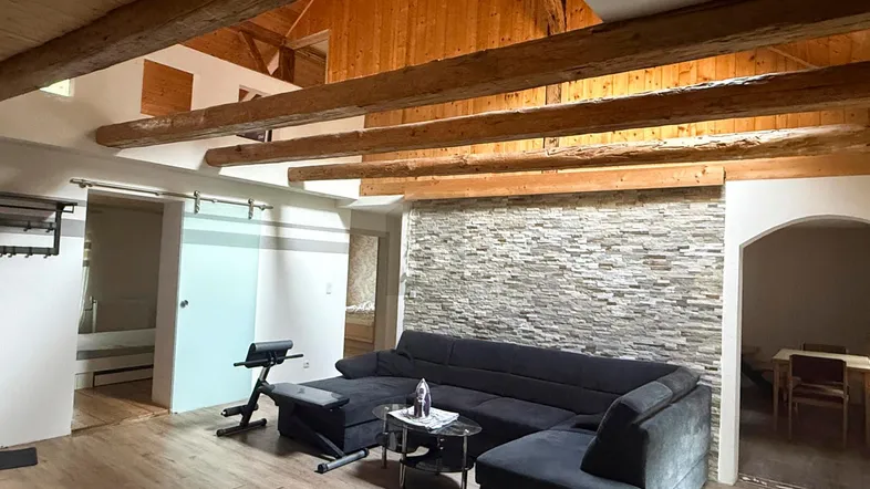 Geräumiges Wohnzimmer mit Holzbalken, Steinwand, dunklem Sofa und offenem Galeriebereich.