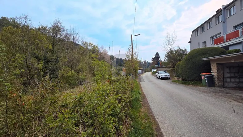 Blick auf eine ländliche Straße mit angrenzendem Gebäude und bewaldetem Hügel.