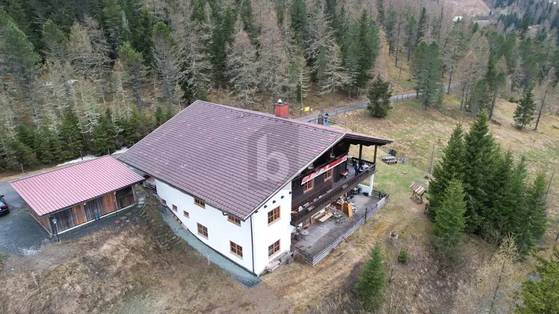 Luftaufnahme eines charmanten Chalets mit Satteldach, Balkonen und Terrasse, umgeben von Wald und Bergen.