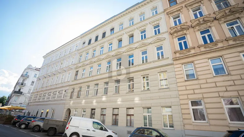 Historische Fassade eines Mehrfamilienhauses mit vielen Fenstern und parkenden Autos auf der Straße.