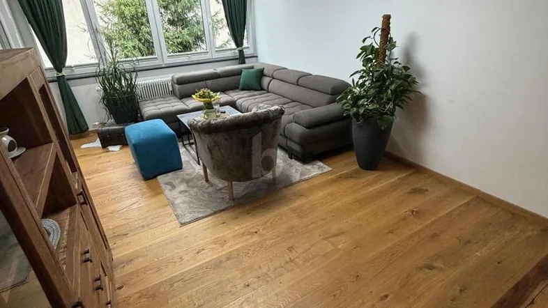 Helles Wohnzimmer mit großem Ecksofa, Holzboden und Blick ins Grüne durch die Fenster.