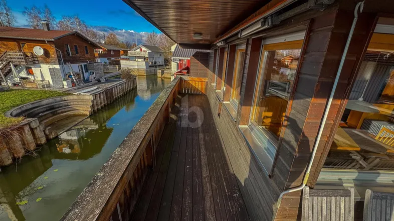 Geräumiger Holzbalkon mit Blick auf den Kanal und die umliegende Landschaft mit Bergen im Hintergrund.