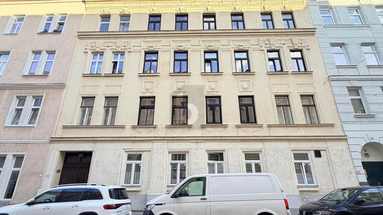 Fassade eines Altbaus mit mehreren Fenstern und geparkten Autos auf der Straße.