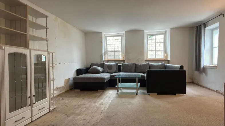 Helles Wohnzimmer mit Ecksofa, Regalen und Teppichboden, der renovierungsbedürftig ist.