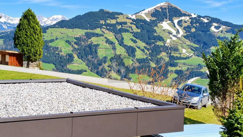 Blick auf die umliegende Berglandschaft mit schneebedeckten Gipfeln und grünen Hängen. Ein Flachdach ist im Vordergrund sichtbar.