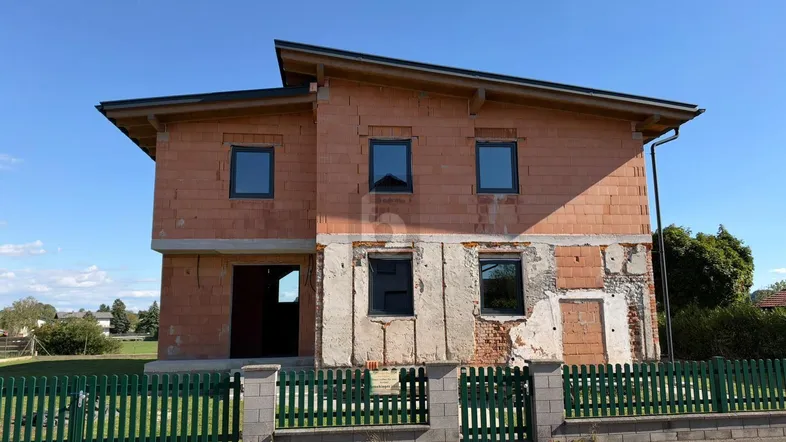 Neubau-Einfamilienhaus im Rohbauzustand mit grüner Umzäunung und blauer Himmel.