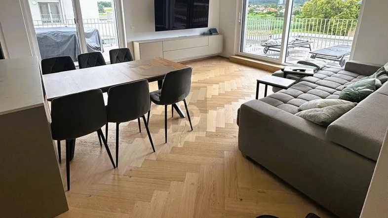 Helles Wohn- und Esszimmer mit Fischgrätparkett, großen Fenstern und Zugang zum Balkon.