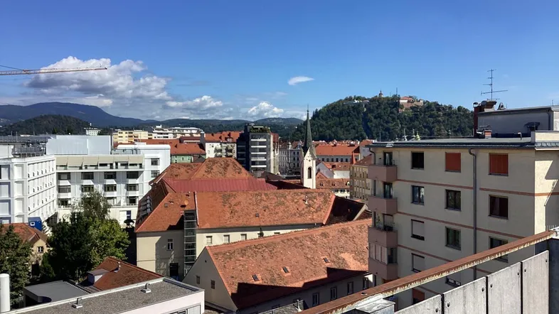 Panoramablick über die Stadt mit roten Dächern und einem Hügel im Hintergrund unter blauem Himmel.