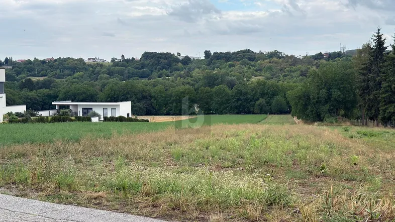 Weites Feld mit modernen Häusern im Hintergrund und grüner Hügellandschaft unter bewölktem Himmel.