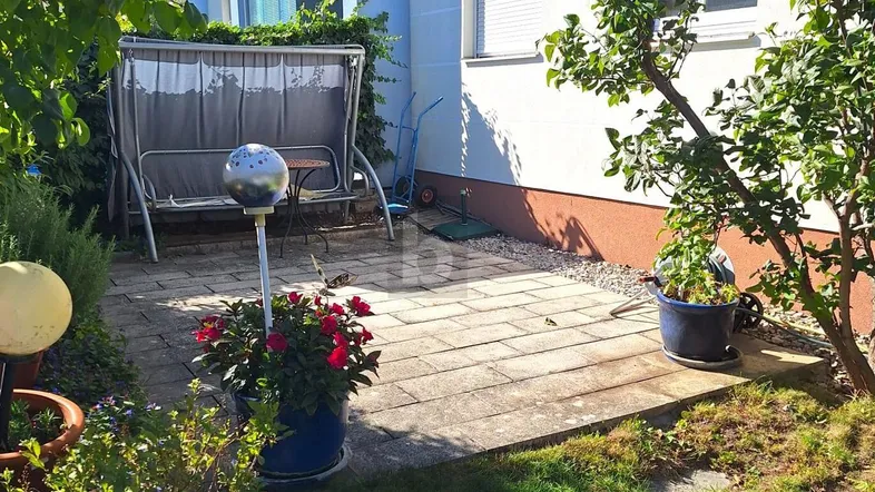 Gepflasterte Terrasse mit Gartenschaukel und blühenden Pflanzen, umgeben von Grünflächen.