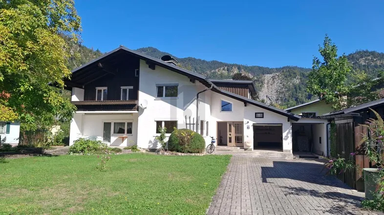 Weißes Haus mit dunklem Dach, gepflegtem Vorgarten und Auffahrt, umgeben von grünen Bergen.