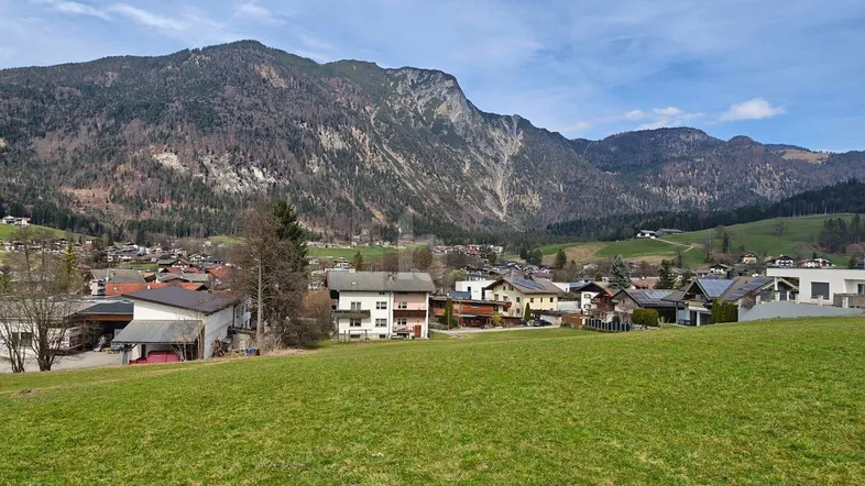 Grüne Wiesen im Vordergrund mit einem Dorf und majestätischen Bergen im Hintergrund unter blauem Himmel.