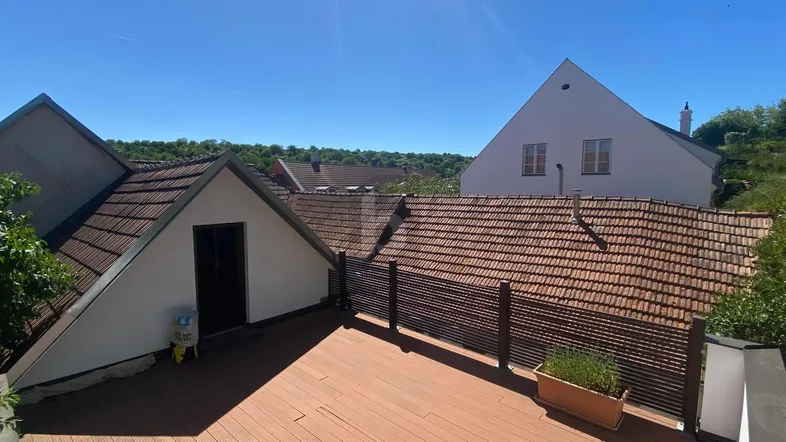 Sonnige Dachterrasse mit Holzboden und Blick auf die umliegenden Dächer und grüne Hügel.