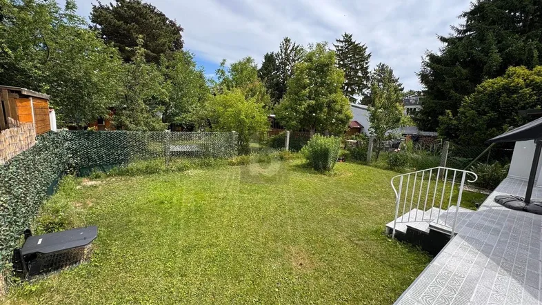Großer Garten mit Rasenfläche, umgeben von Bäumen und Sträuchern, zugänglich über eine kleine Treppe.