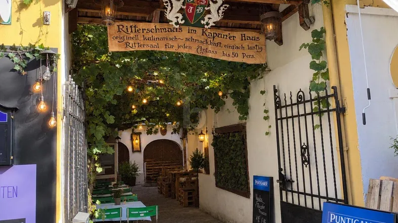 Eingangsbereich eines Restaurants mit üppigem Grün, Lichterketten und einem traditionellen Holzschild.