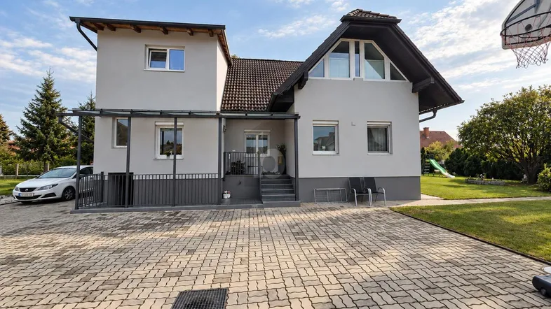 Modernes Einfamilienhaus mit gepflasterter Auffahrt, Gartenbereich und Basketballkorb unter blauem Himmel.