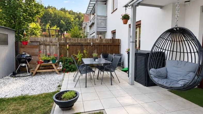 Gepflasterte Terrasse mit Gartenmöbeln, Hängesessel und Grillbereich, umgeben von Grün.