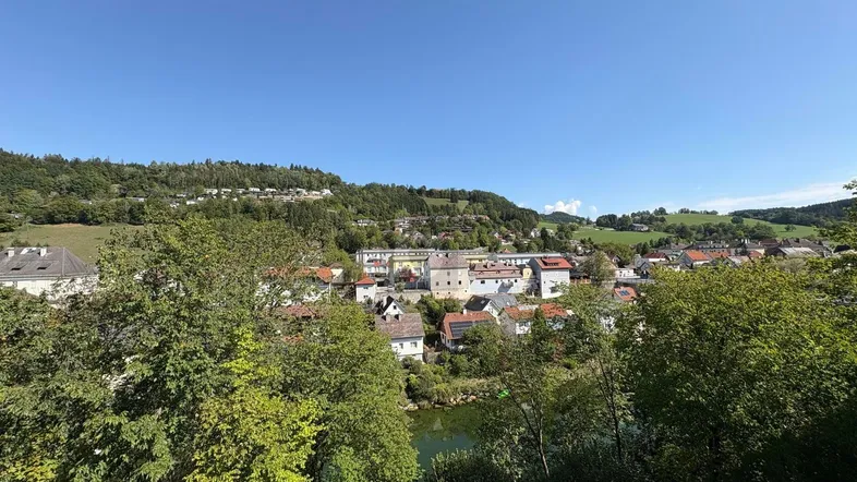 Panoramablick über eine malerische Stadt im Tal mit Fluss und grünen Hügeln unter blauem Himmel.