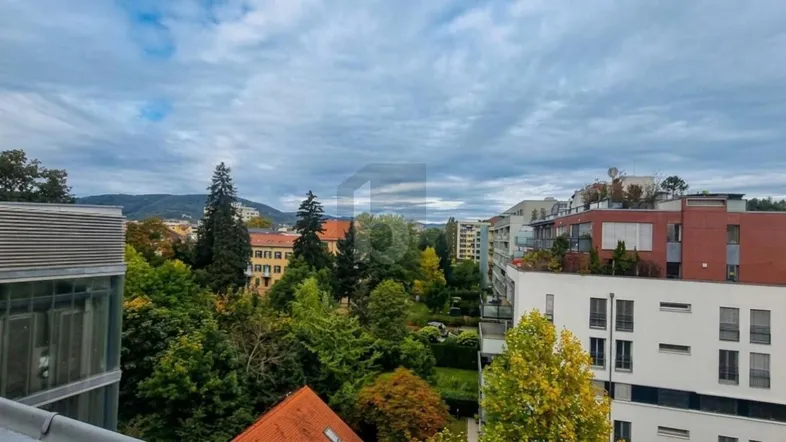 Panoramablick über die Stadt mit grünen Bäumen und Bergen im Hintergrund unter bewölktem Himmel.