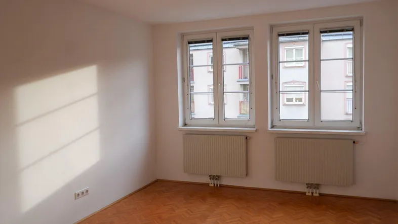 Helles Zimmer mit Fischgrätparkett und zwei Fenstern, die Tageslicht hereinlassen.