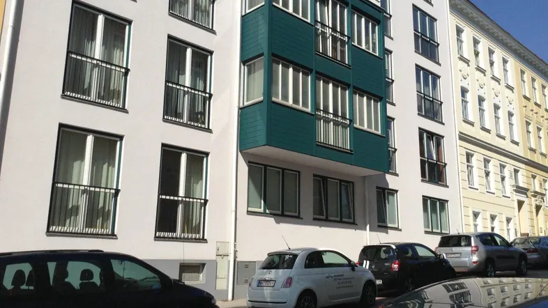 Moderne Wohnhausfassade mit markantem türkisfarbenem Erker und vielen Fenstern an einer städtischen Straße.