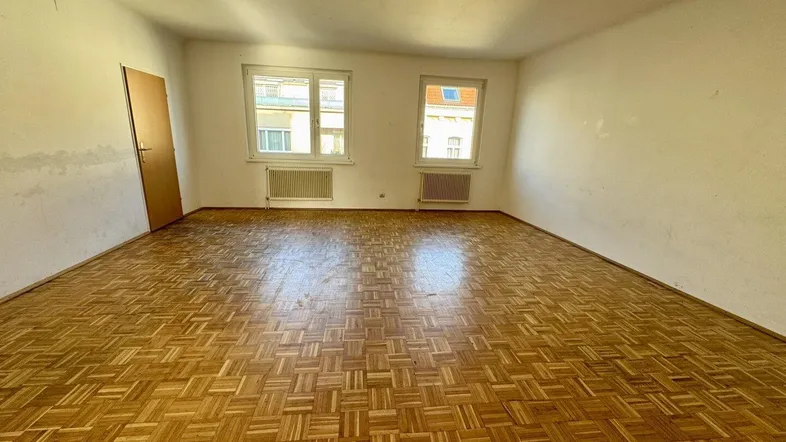 Helles Wohnzimmer mit klassischem Parkettboden und zwei Fenstern, die viel Tageslicht hereinlassen.