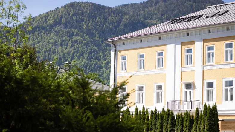 Gelbe Fassade eines Gebäudes mit weißen Fenstern und Blick auf bewaldete Berge.