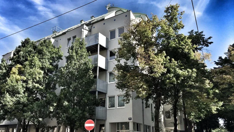 Modernes Mehrfamilienhaus mit Balkonen und Bäumen an einer städtischen Straße.