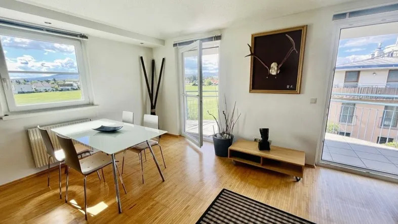 Helles Wohn- und Esszimmer mit Holzboden, Zugang zum Balkon und weitem Blick ins Grüne.