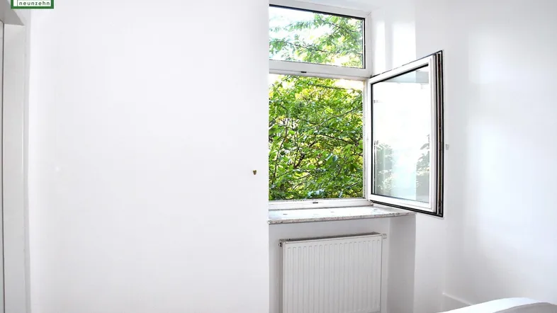 Helles Schlafzimmer mit Fensterblick ins Grüne und einem Heizkörper unter dem Fenster.