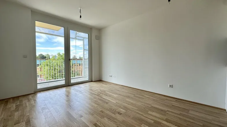Helles, leeres Zimmer mit Holzboden und Zugang zu einem Balkon, ideal für einen Wohnbereich.
