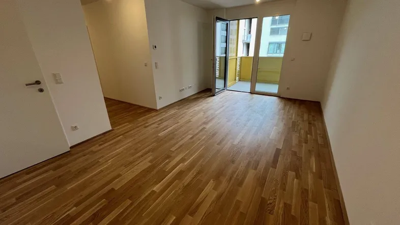 Helles, leeres Wohnzimmer mit Parkettboden und Zugang zu einem Balkon.