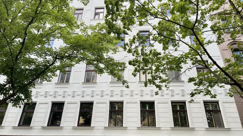 Weiße Fassade eines Mehrfamilienhauses mit klassischen Verzierungen und grünen Bäumen im Vordergrund.