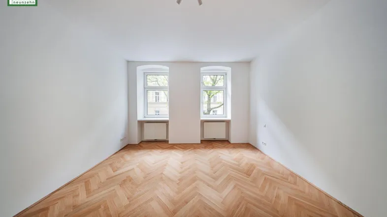 Helles, leeres Zimmer mit Fischgrätparkettboden und zwei Fenstern, die viel Tageslicht hereinlassen.