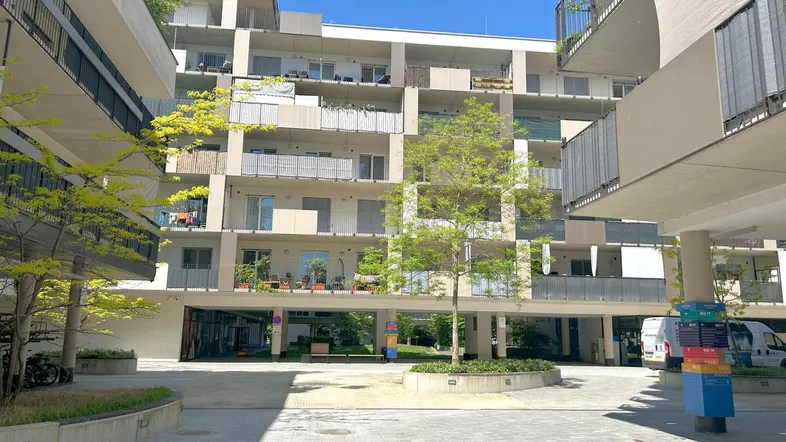 Moderner Apartmentkomplex mit mehreren Balkonen und einem begrünten Innenhof unter blauem Himmel.