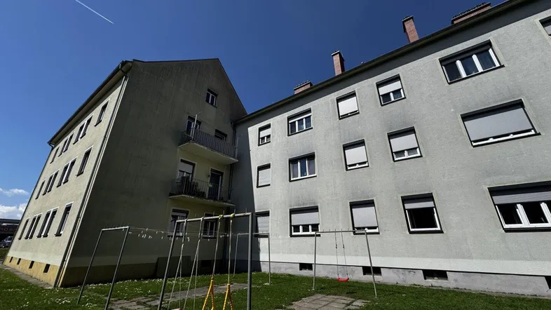Mehrfamilienhaus mit hellgrüner Fassade und kleinem Spielplatz im Vorgarten unter blauem Himmel.