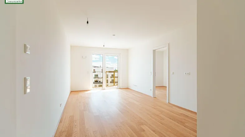 Lichtdurchfluteter Raum mit Holzboden und großen Fenstern, die Zugang zu einem Balkon bieten.
