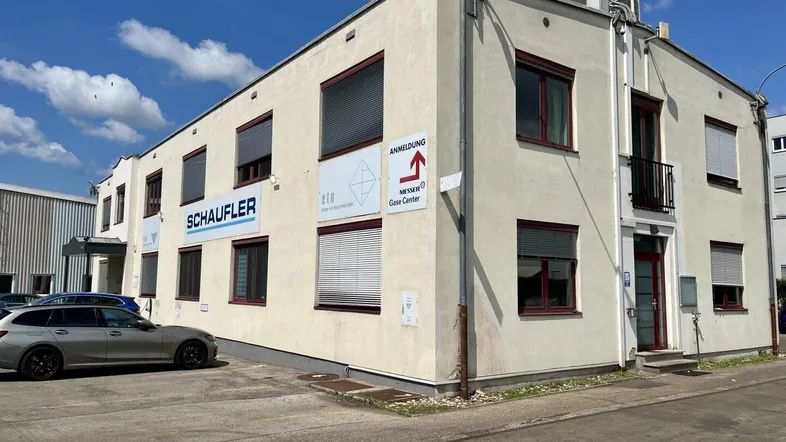 Das Gebäude präsentiert sich mit einer hellen Fassade, roten Fensterrahmen und einem Parkplatz davor.