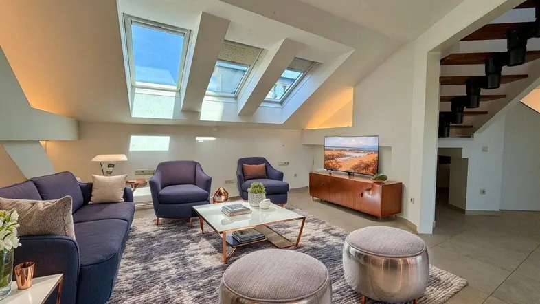 Helles Wohnzimmer im Dachgeschoss mit gemütlichen Möbeln, Dachfenstern und sichtbarer Treppe.