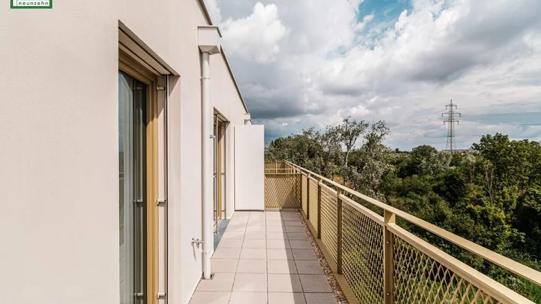 Langer Balkon mit Fliesenboden und Blick ins Grüne unter bewölktem Himmel.