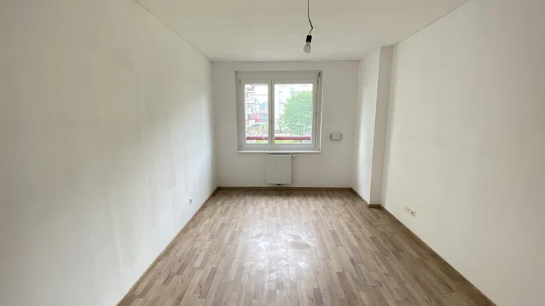 Helles, leeres Zimmer mit Parkettboden und einem Fenster, das Tageslicht hereinlässt.