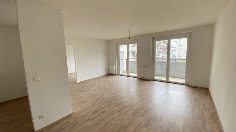 Helles, leeres Wohnzimmer mit Laminatboden und Zugang zu einem Balkon.