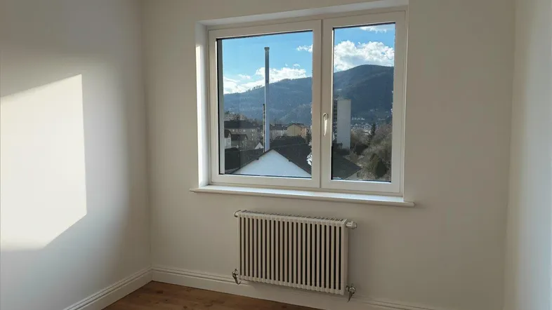 Helles Zimmer mit Holzboden, Heizkörper und weitem Blick auf die umliegende Berglandschaft.