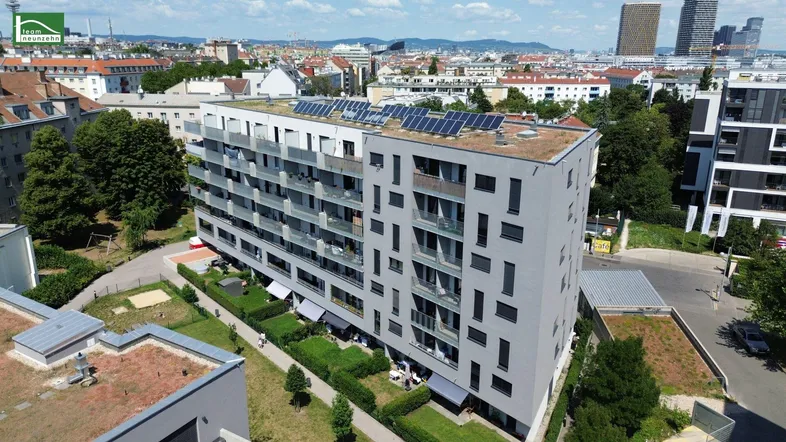Luftaufnahme eines modernen Mehrfamilienhauses mit Solaranlage auf dem Dach und städtischer Umgebung.