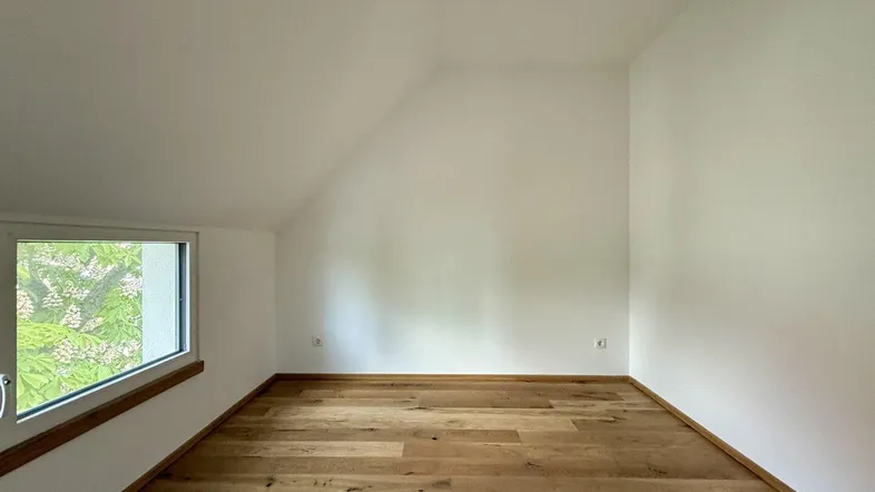 Helles Zimmer im Dachgeschoss mit Holzboden und schrägem Fenster, bereit zur Einrichtung.