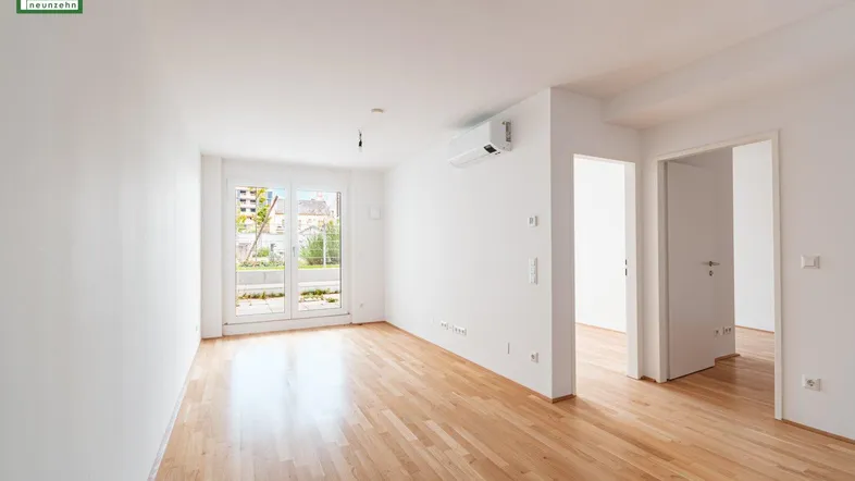 Helles, leeres Wohnzimmer mit Holzboden, weißen Wänden und Zugang zu einem Außenbereich.