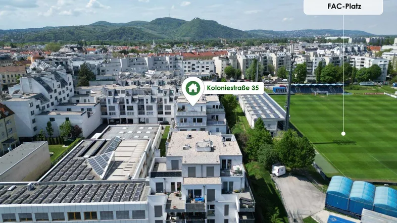 Luftaufnahme einer Wohnanlage mit umliegenden Gebäuden und grünen Hügeln im Hintergrund.