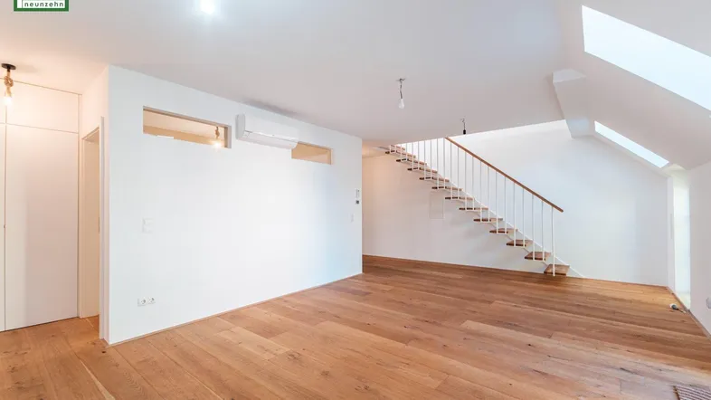 Geräumiger Wohnbereich mit Holzboden, weiß gestrichenen Wänden und offener Treppe zum Obergeschoss.