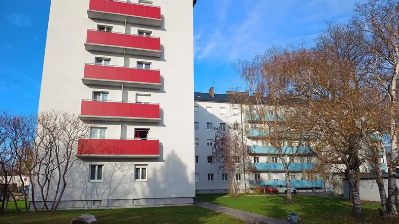 Mehrfamilienhaus mit roten Balkonen und Grünflächen in einer Wohnsiedlung unter blauem Himmel.